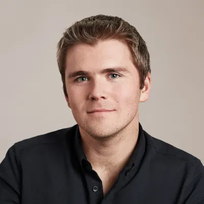 Headshot of Stripe co-founder and President John Collison, wearing a dark collared shirt against a light gray background.